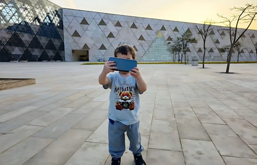 A Young Tourist Child stands outside the modern, geometric facade of The Grand Egyptian Museum (GEM) and is Taking Selfie with a smartphone.