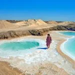 A woman in a pink dress walks along the brilliant white edge of the Salt Ponds Of Lake Aghurmi in the Siwa Oasis, contrasting with the turquoise water and surrounding desert landscape.