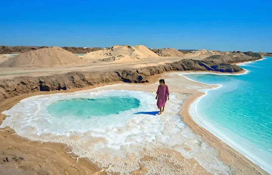 A woman in a pink dress walks along the brilliant white edge of the Salt Ponds Of Lake Aghurmi in the Siwa Oasis, contrasting with the turquoise water and surrounding desert landscape.