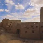 An atmospheric, winding, dusty alleyway lined by the crumbling mud-brick of the Sandstone Walls and Ancient Fortress of an Old Shali Mountain village in Siwa Oasis, with a towering conical structure in the distance.