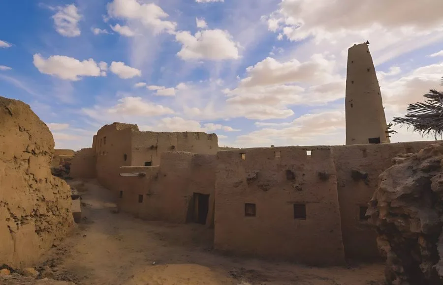 An atmospheric, winding, dusty alleyway lined by the crumbling mud-brick of the Sandstone Walls and Ancient Fortress of an Old Shali Mountain village in Siwa Oasis, with a towering conical structure in the distance.