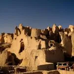 A close-up view of the Sandstone Walls and Ancient Fortress of an Old Shali Mountain village in Siwa Oasis, showing the towering, rugged mud-brick and salt ruins under a clear blue sky.