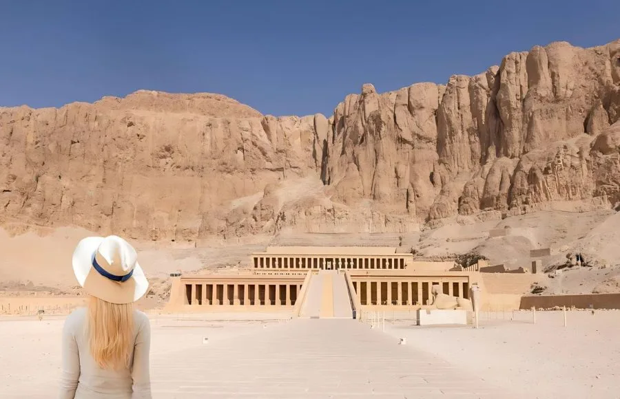 A blonde haired woman in a hat stands in awe of the stunning Mortuary Temple of Hatshepsut in Luxor, Egypt