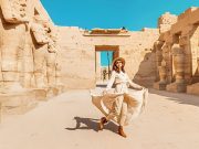 A happy tourist girl in a dress is interested at the Karnak Temple, posing amidst the ancient statues and sunlit stone walls.