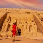A young tourist girl in red dress walking towards the Abu Simbel Temple, highlighting the colossal statues carved into the sandstone cliff face.