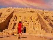 A young tourist girl in red dress walking towards the Abu Simbel Temple, highlighting the colossal statues carved into the sandstone cliff face.
