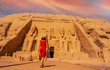 A young tourist girl in red dress walking towards the Abu Simbel Temple, highlighting the colossal statues carved into the sandstone cliff face.