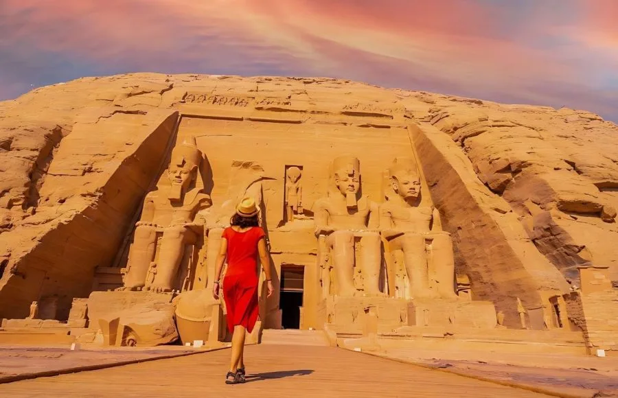 A young tourist girl in red dress walking towards the Abu Simbel Temple, highlighting the colossal statues carved into the sandstone cliff face.