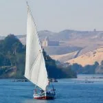 Felucca-Glides-Along-The-River-Nile-Aswan-Egypt-With-Aga-Khan-On-The-Hill-In-The-Distance