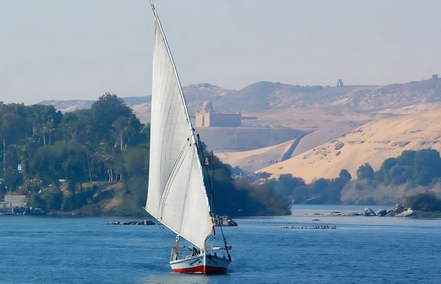 Felucca-Glides-Along-The-River-Nile-Aswan-Egypt-With-Aga-Khan-On-The-Hill-In-The-Distance