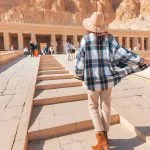 A tourist in a wide-brimmed hat and flowing shirt walking up the steps towards the majestic Hatshepsut Temple.