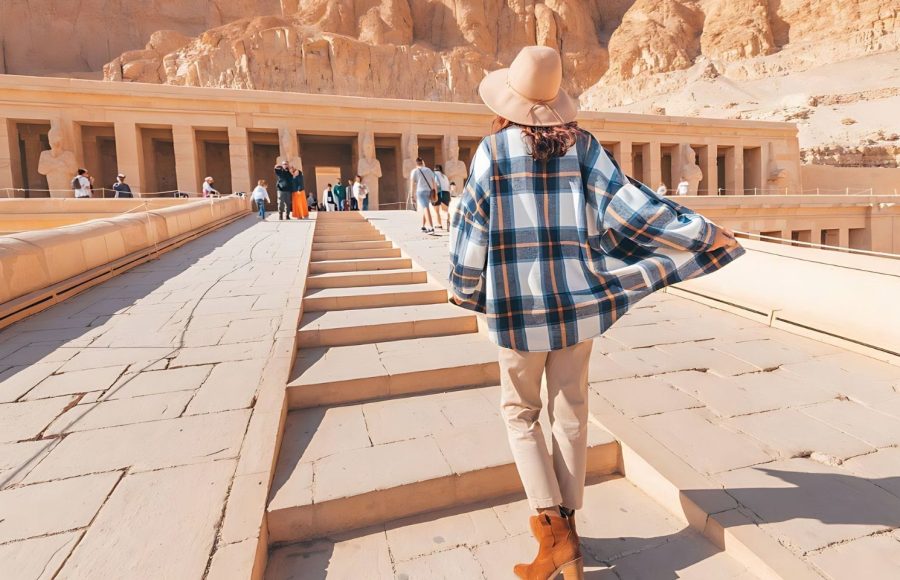 A tourist in a wide-brimmed hat and flowing shirt walking up the steps towards the majestic Hatshepsut Temple.