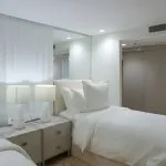 Interior view of a twin-bed cabin on the Kazazian-Berge Dahabiya Nile cruise boat, showing two beds with white linens, a nightstand, and a view into an adjacent bathroom area.