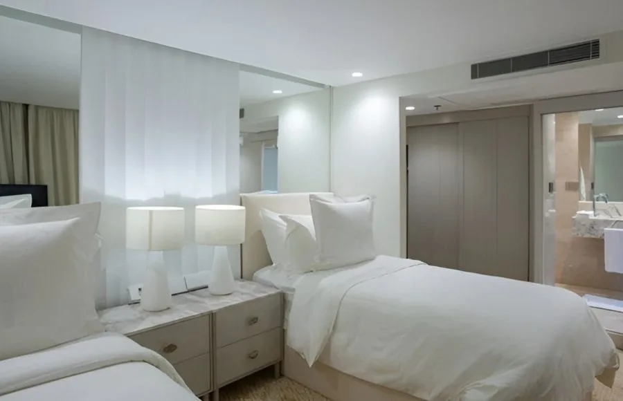 Interior view of a twin-bed cabin on the Kazazian-Berge Dahabiya Nile cruise boat, showing two beds with white linens, a nightstand, and a view into an adjacent bathroom area.
