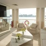 The elegant, light-filled lounge area of the Kazazian Berge luxury motor sailer yacht, featuring white sofas and large windows overlooking the Nile River.