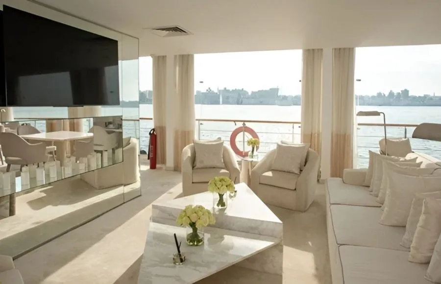 The elegant, light-filled lounge area of the Kazazian Berge luxury motor sailer yacht, featuring white sofas and large windows overlooking the Nile River.