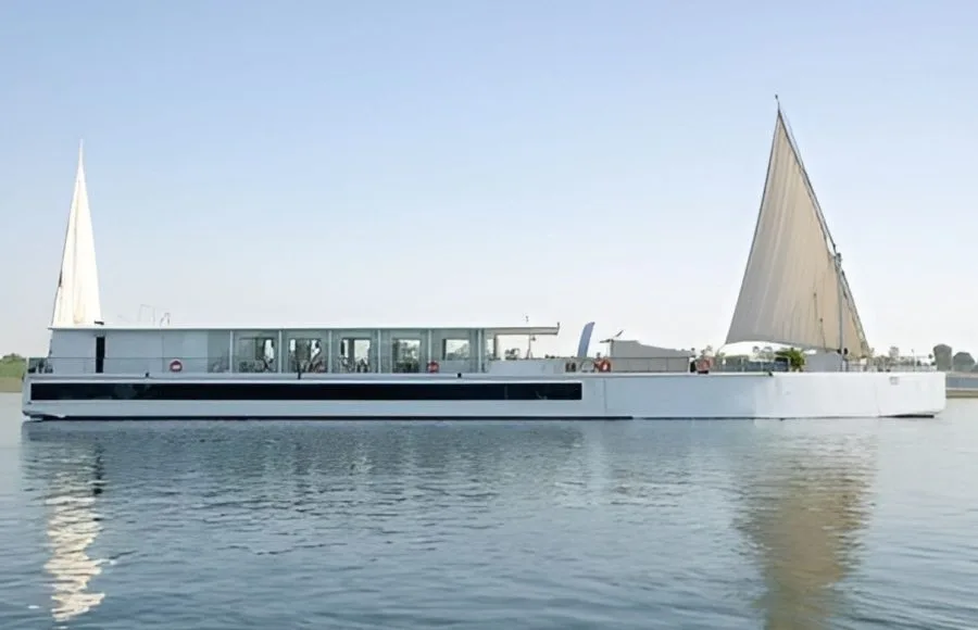 A white luxury Dahabiya sailing vessel with large sails on the Nile River, surrounded by water and lush green riverbanks in Egypt.