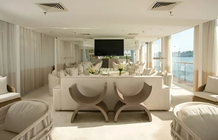 A luxurious lounge area on the Kazazian Berge Dahabiya yacht with white sofas, chairs, large windows overlooking the Nile, and a flat-screen TV.