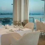 A dining room on a luxury Nile cruise ship with set tables, modern chairs, and large windows overlooking the river and the opposite bank of the Nile.