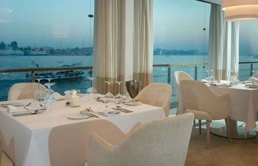 A dining room on a luxury Nile cruise ship with set tables, modern chairs, and large windows overlooking the river and the opposite bank of the Nile.