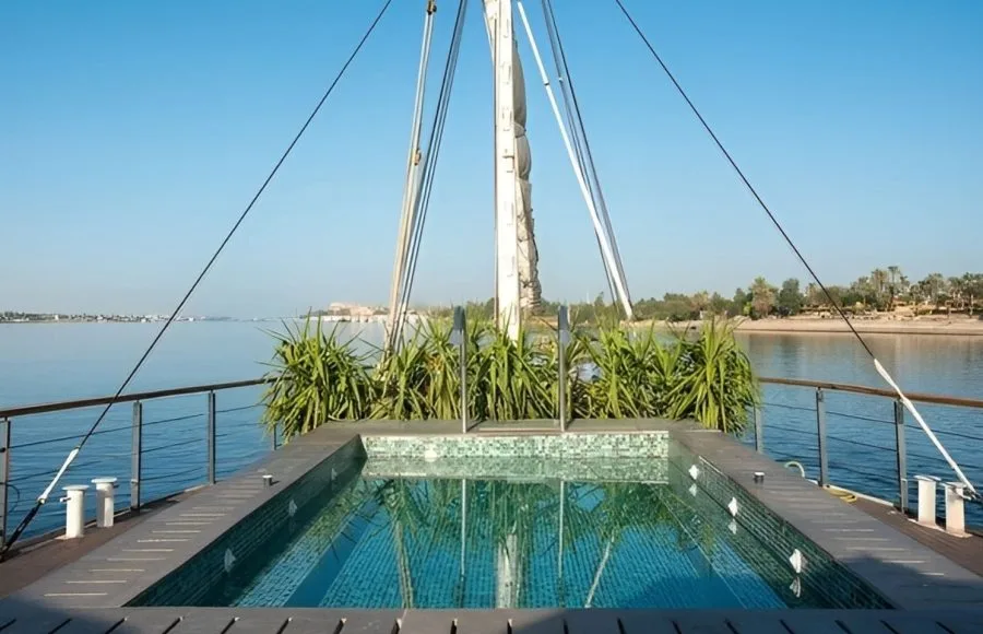 Sun deck of the Kazazian Berge Dahabiya, a luxury Nile cruise ship, featuring a rectangular plunge pool with jets and lush green riverbanks in the background.