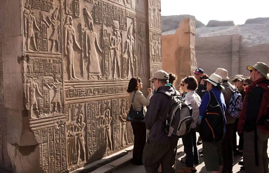 A group of tourists and a guide studying the detailed relief carvings on the wall of the Kom Ombo Temple of Horus and Sobek, illustrating ancient Egyptian figures and hieroglyphs.