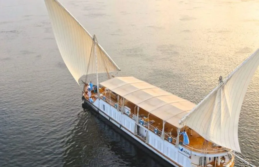 A traditional two-masted Egyptian sailboat, a Lazuli Dahabiya, cruising on the wide Nile River under a clear blue sky.