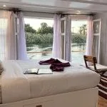 A view inside a luxury cabin on the Lazuli Abydos Dahabiya Nile Cruise boat, showing a large bed, open French doors revealing a river view, and small furniture.