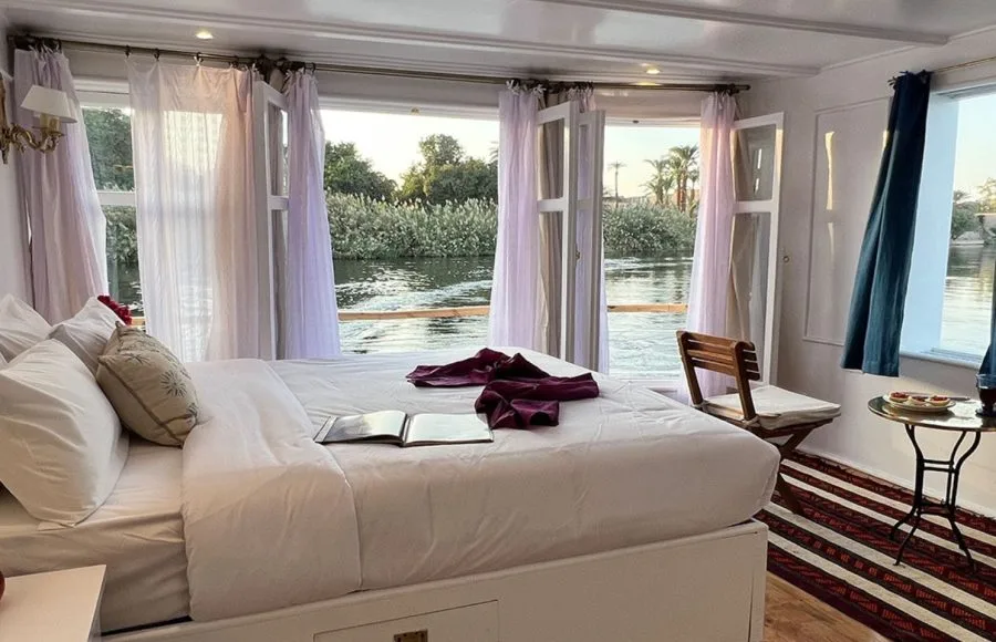 A view inside a luxury cabin on the Lazuli Abydos Dahabiya Nile Cruise boat, showing a large bed, open French doors revealing a river view, and small furniture.