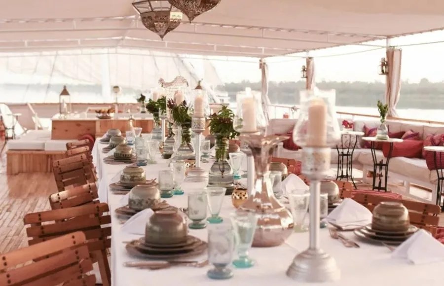 A dining table on the upper deck of the Lazuli Abydos Dahabiya Nile Cruise boat, set with fine china and silverware, ready for a meal with a river view.