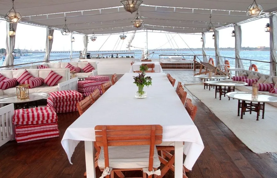 Sun deck of the Lazuli Abydos Dahabiya Nile Cruise boat, featuring a set dining table, lounge chairs, and views of the Nile.