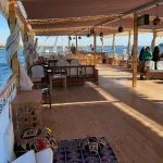 A spacious wooden sundeck on a cruise boat with lounge seating, patterned floor cushions, and an overhead canopy overlooking the Nile River.