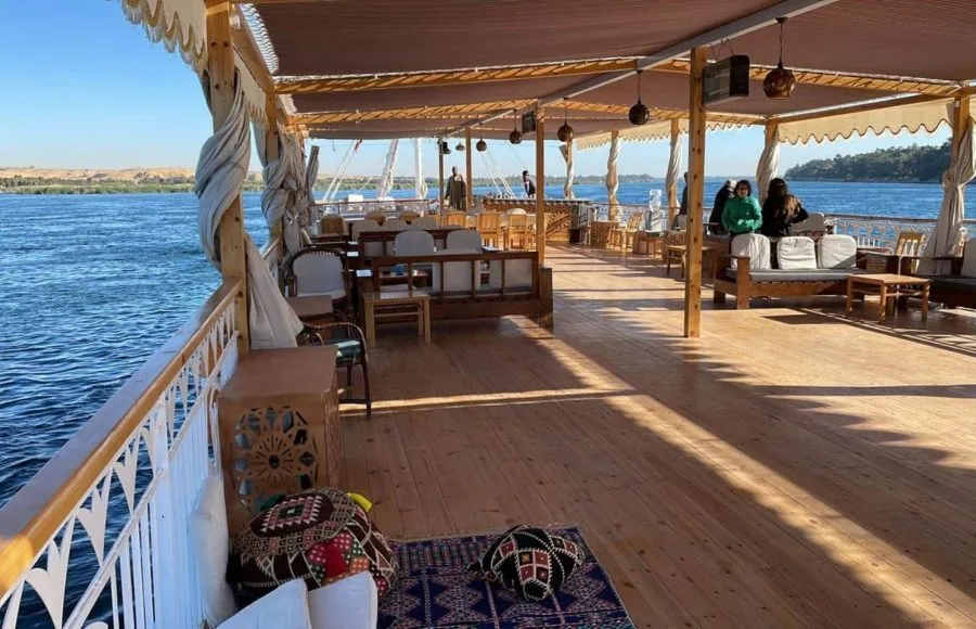 A spacious wooden sundeck on a cruise boat with lounge seating, patterned floor cushions, and an overhead canopy overlooking the Nile River.