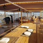 A spacious wooden boat sundeck featuring floor cushions on rugs, wooden benches with white pillows, and a shaded canopy overlooking a river and bridge.