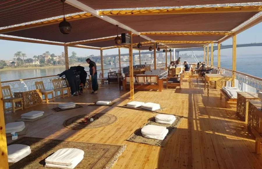 A spacious wooden boat sundeck featuring floor cushions on rugs, wooden benches with white pillows, and a shaded canopy overlooking a river and bridge.