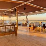 An outdoor lounge area on a wooden boat deck with wooden sofas, white cushions, and small coffee tables under a striped canopy during sunset.