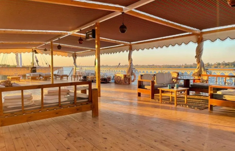 An outdoor lounge area on a wooden boat deck with wooden sofas, white cushions, and small coffee tables under a striped canopy during sunset.