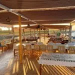 A long dining table with white chairs set under a canopy on the wooden deck of a boat at sunset.