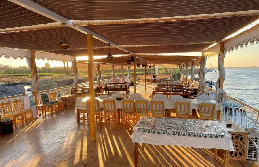 A long dining table with white chairs set under a canopy on the wooden deck of a boat at sunset.