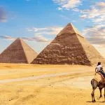 A tourist rides a camel guided by a bedouin in the Giza desert, with the colossal The Egypt Pyramids Khafre and Cheops rising dramatically in the background under a vivid sky.