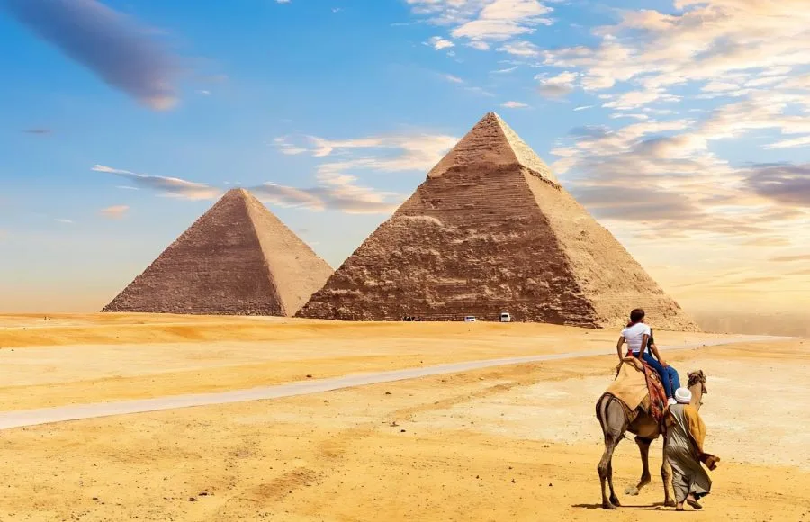 A tourist rides a camel guided by a bedouin in the Giza desert, with the colossal The Egypt Pyramids Khafre and Cheops rising dramatically in the background under a vivid sky.