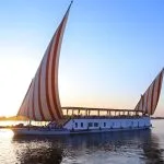 A traditional Egyptian dahabiya sailboat with red and white striped sails glides on the Nile River at sunset.