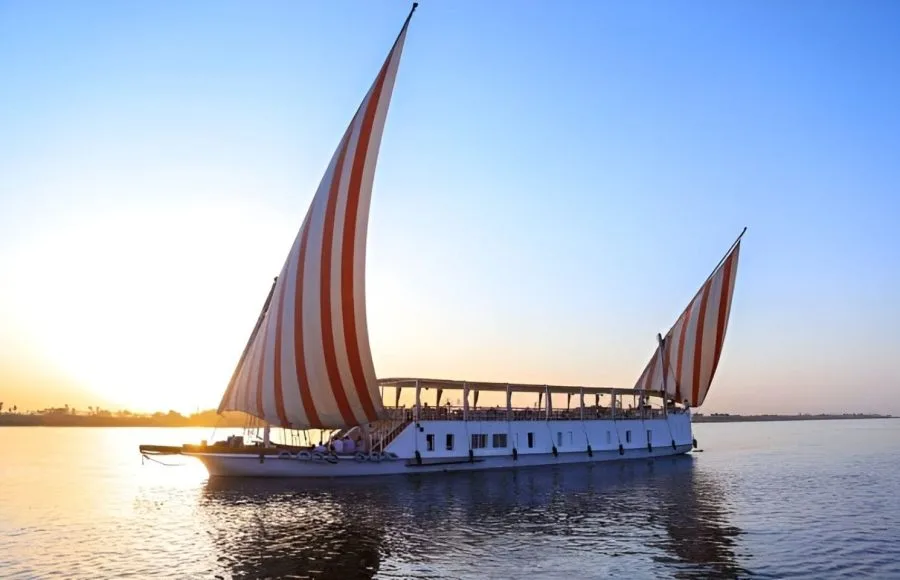 A traditional Egyptian dahabiya sailboat with red and white striped sails glides on the Nile River at sunset.