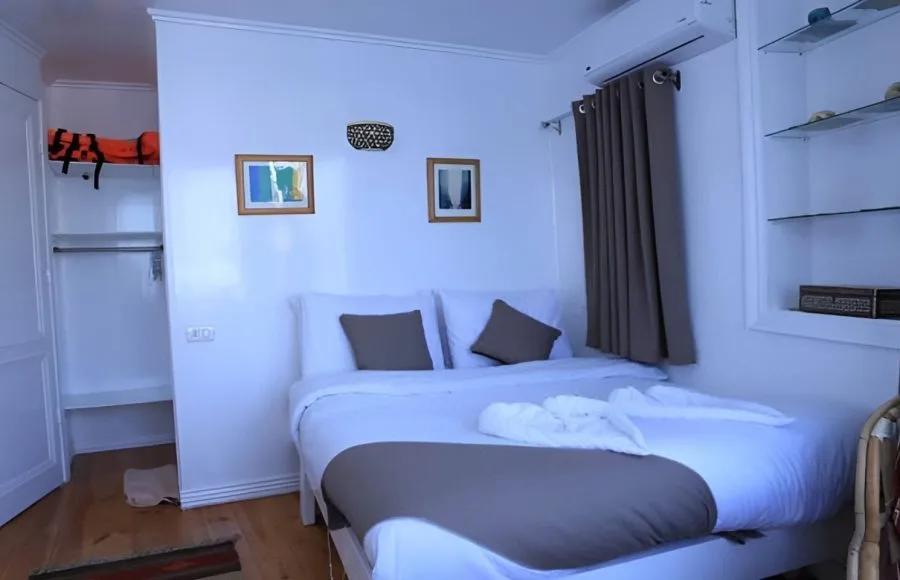 Interior view of a cozy, air-conditioned cabin on a traditional Egyptian Dahabiya sailboat with a bed and a view of the Nile River through a large window.