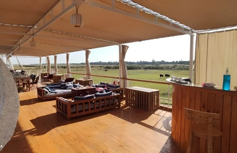 A traditional Egyptian Dahabiya sailing boat deck, the "Three Pyramids Dahabiya", featuring a partially shaded sun deck with oriental seating, wooden lounges, a small bar, and panoramic views of the green Nile riverbanks and fields during a cruise.