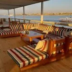 The deck of a Dahabiya Nile Cruise boat with striped seating, a table, and a view of the river.