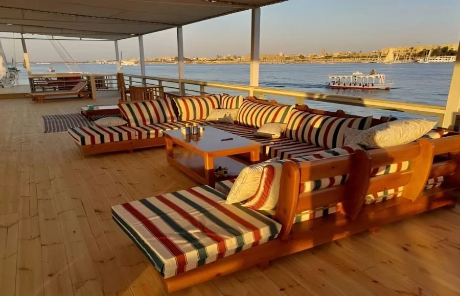 The deck of a Dahabiya Nile Cruise boat with striped seating, a table, and a view of the river.