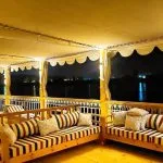 Deck of the "Three Pyramids" Dahabiya Nile Cruise at night, featuring a long, shaded seating area with wooden bench sofas and striped pillows on both sides, overlooking the dark river and distant shore lights.