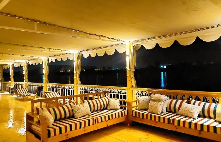 Deck of the "Three Pyramids" Dahabiya Nile Cruise at night, featuring a long, shaded seating area with wooden bench sofas and striped pillows on both sides, overlooking the dark river and distant shore lights.
