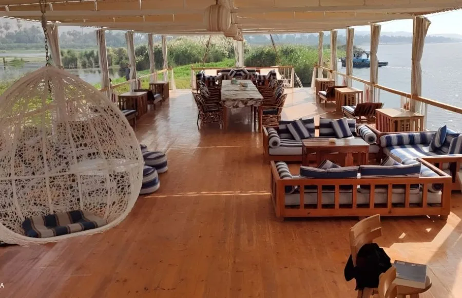 The sundeck of a traditional Egyptian Dahabiya cruise boat on the Nile River, featuring white wicker hanging chairs, wooden seating areas with blue and white striped cushions, dining tables set for a meal, and a view of the river bank and another boat in the distance. The website interface for "Tripidays Egypt Tours" is partially visible.